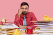 Lazy displeased fatigue male worker keeps fist on cheek, has sleepy look, dressed in formal shirt and tie, works with papers, has mess on workpace, poses over pink background, being overload