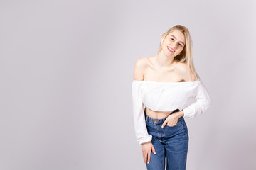 Studio shot of gorgeous young blonde woman with straight hair wearing off shoulder crop top sweater and high waisted denim shorts. Gray isolated background, copy space, close up.