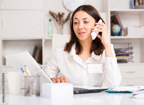 Fotografie, Obraz Young female using laptop and talking by smartphone at the office