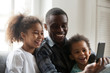 Happy african american dad laughing using smartphone with little son daughter at home, black daddy and children make call selfie watching funny video on phone, family father and cute kids having fun