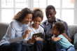 © fizkes - Black african family of mom dad parents and happy 2 mixed race little kids sit together on sofa laughing looking at smartphone taking selfie, making video call, watching funny video on phone at home