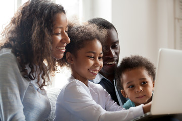 Wall Mural - Happy black family with kids having fun using laptop application together at home, african parents and children laughing doing shopping, watching funny internet video, making online computer call