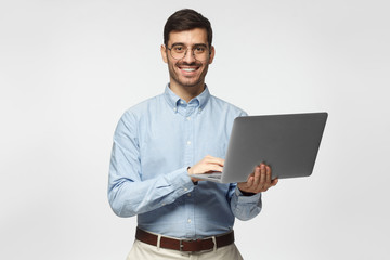 Wall Mural - Handsome young man using laptop and looking at camera with smile, isolated on gray background