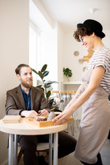 Wall Mural - Smiling friendly waitress in hat bringing order to customer in modern coffee shop and putting tray on table, cheerful bearded man in earphones smiling at waitress