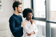 © alfa27 - Positive african woman embraces with her european handsome brunette boyfriend, relaxing at home, give warm hug to each other, stand near window background. Multiethnic relationship and love concept.