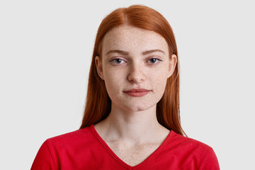 Headshot of attractive red haired European woman with freckled skin, looks seriously at camera, has minimal make up, wears red sweater, isolated over white background. Natural beauty concept