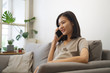 © Kawee - Happy young Asian woman using mobile phone talking with her friend with smile while sitting on a couch at home in the living room. Technology and lifestyle concept.
