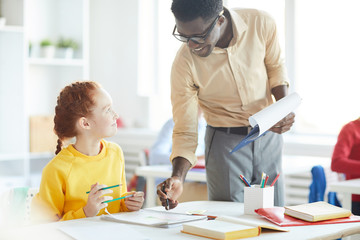 young teacher consulting little girl while pointing at her notes in copybook at lesson in classroom