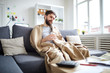 © pressmaster - Sick man wrapped into blanket sitting on sofa in front of table with papers while staying at home
