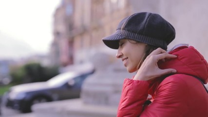 Wall Mural - Portrait of a young smiling woman in red jacket and grey cap. Woman sitting outdoors on the street at sunny autumn or spring day and fasten jacket