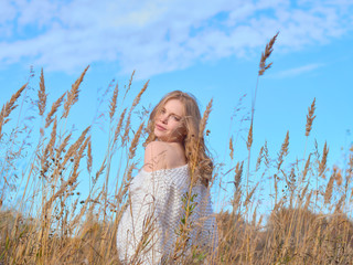 Beautiful pregnant woman in a white woolen sweater. Beautiful pregnant woman in high dry grass against the blue sky.