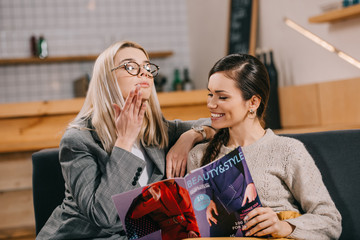 attractive woman looking at friend while holding beauty magazine