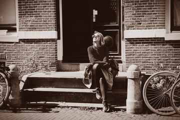 Wall Mural - Young girl in sunglasses and sweater with backpack sitting at street in Amsterdam. Holland, Netherlands. Autumn season Image in sepia color style