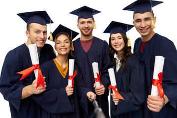 Wall Mural - education, graduation and people concept - group of happy graduate students in mortar boards and bachelor gowns with diplomas taking picture by slfie stick over white background