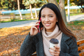 Sticker - Smiling young asian woman wearing coat sitting on a bench