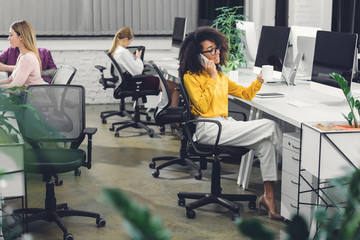 Wall Mural - african american businesswoman holding cup and talking by smartphone while working with colleagues in office