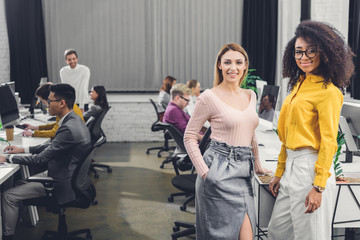 Wall Mural - young multiethnic businesswomen smiling at camera while colleagues working behind in office