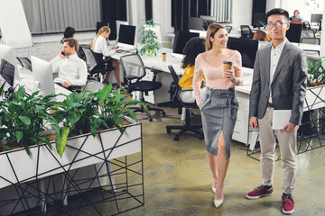 Wall Mural - smiling young businesswoman holding coffee to go and looking at happy asian colleague with digital tablet in open space office