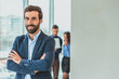 © Andrii - A businessman stands in the office folded his hand to his hand. During this time, he stands against the background of his colleagues who work in the laptop.