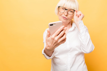 Selfie time! Positive aged woman smiling while using phone isolated over yellow background.