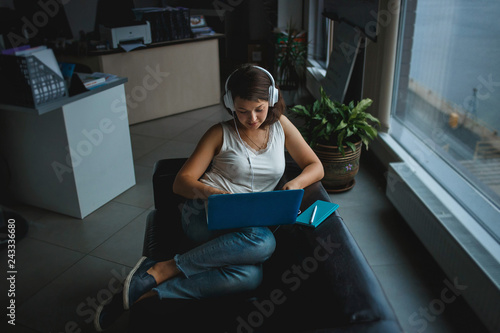 Fotografie, Obraz Young woman listening music in headphones and working on laptop on the sofa in office