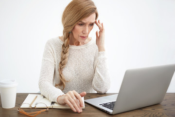 Stressed successful elderly businesswoman in white sweater touching forehead, having headache while working online using laptop, sitting at table with copybook, drinking another cup of coffee