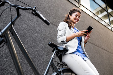 Wall Mural - Young businesswoman standing on a city street with bicycle looking at phone. 