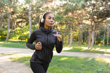 Image of energetic woman 20s wearing black tracksuit and headphones working out, while running through green park