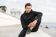 © Drobot Dean - Image of sportive adult man 30s in black sportswear, sitting on boardwalk at seaside