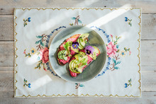 Top Shot Of Two Slices Avocado Rye Toast On Beetroot Hummus With Edible Purple Pansy Flowers, Micro Greens And Nigella Seeds Or Onion Seeds, On A Blue Plate, On Embroided Teatowel On White Wooden Back