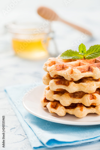 Fotografía Belgian waffles with mint leaves and honey in glass jar with wooden honey dipper on pastel blue background