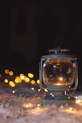 Lantern with burning candle and Christmas lights on white snow outdoors