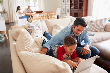 Wall Mural - Pre-teen boy lying on sofa using laptop with his dad. Mum and grandad at the table in the background