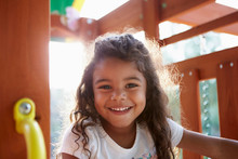 Young Hispanic Girl Playing On A Climbing Frame In A Playground Smiling To Camera, Backlit, Close Up