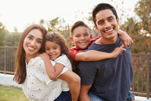 Young Hispanic Parents Piggyback Their Children In The Park, Smiling To Camera, Focus On Foreground