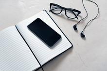 An Open Notebook With A Black Cover On It Is A Black Mobile Phone, Next To It On A White Background Are Glasses With Black Rim And Black Wired Music Headphones, There Is An Empty Space For A Signature