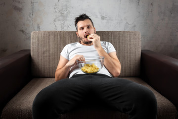 Wall Mural - The guy in the shirt is lying on the couch, eating chips and watching a sports channel. The concept of laziness, frustration, procrastination, the person at home.