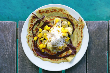 Wall Mural - A tropical breakfast crepe with pineapple, chocolate, and fresh coconut eaten by the blue sea