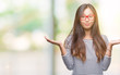 Young asian woman wearing glasses over isolated background clueless and confused expression with arms and hands raised. Doubt concept.