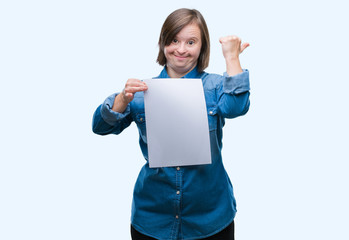 Sticker - Young adult woman with down syndrome holding blank paper sheet over isolated background pointing and showing with thumb up to the side with happy face smiling