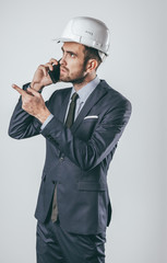 Wall Mural - Handsome guy in white hardhat and elegant suit looking away and pointing aside while standing on light gray background and having smartphone conversation