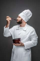 Sticker - Bearded guy in chef uniform keeping eyes closed and trying yummy food from small saucepan while standing on gray background