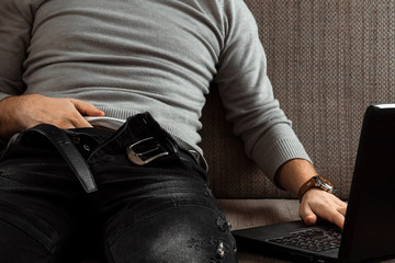 A man watches an adult video on a laptop while sitting on the couch. The concept of porn, masturbation, male needs, pervert, lust, desire, loneliness.