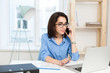 © Look! - A young brunette girl is working at the table in office. She wears blue shirt and black glasses. She is speaking on phone.