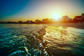 Canvas Print - Golden waves of the sea and city on horizon during sunset