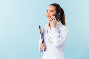 Sticker - Woman doctor posing isolated over blue wall background holding clipboard talking by mobile phone.