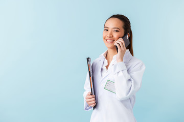 Sticker - Woman doctor posing isolated over blue wall background holding clipboard talking by mobile phone.