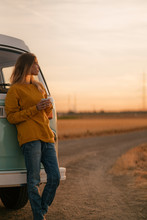 Young Woman With Coffee Cup Standing By Campervan During Sunset