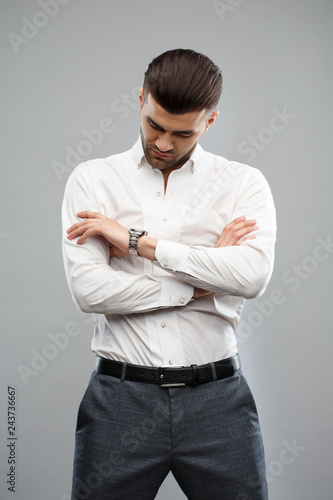 Fotografía Elegant man in casual business clothes standing with arms folded and looking at his watch isolated on a gray background