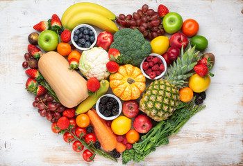 Heart made from healthy rainbow fruits vegetables berries, strawberries oranges plums grapes broccoli cauiliflower mango persimmon pineapple on wooden table, top view, copy space, selective focus